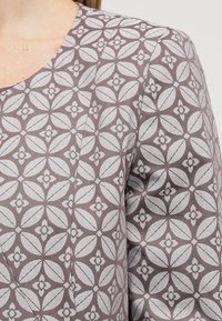 Zbliżenie na tkaninę z powtarzającym się geometrycznym wzorem kwiatowym w białe płatki na stonowanym purpurowym tle.