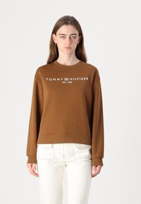 Hnědá mikina s dlouhým rukávem a žebrovanými manžetami a spodním lemem. Na přední straně je logo "TOMMY HILFIGER EST. 1985" v bílé barvě. Spárovaná s bílými kalhotami.