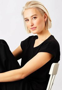 Top corto nero a maniche corte con scollatura a V, indossato da una modella con capelli platino biondi, seduta su una sedia bianca, in posa informale.