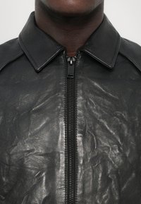 Black leather jacket with a classic collar, featuring a central zipper and slightly wrinkled texture, highlighting its supple material.