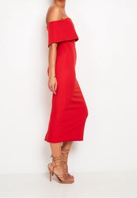 Robe midi rouge à épaules dénudées avec une silhouette ajustée, une texture lisse et un ourlet en dessous du genou. Associée à des talons hauts nus à lanières.