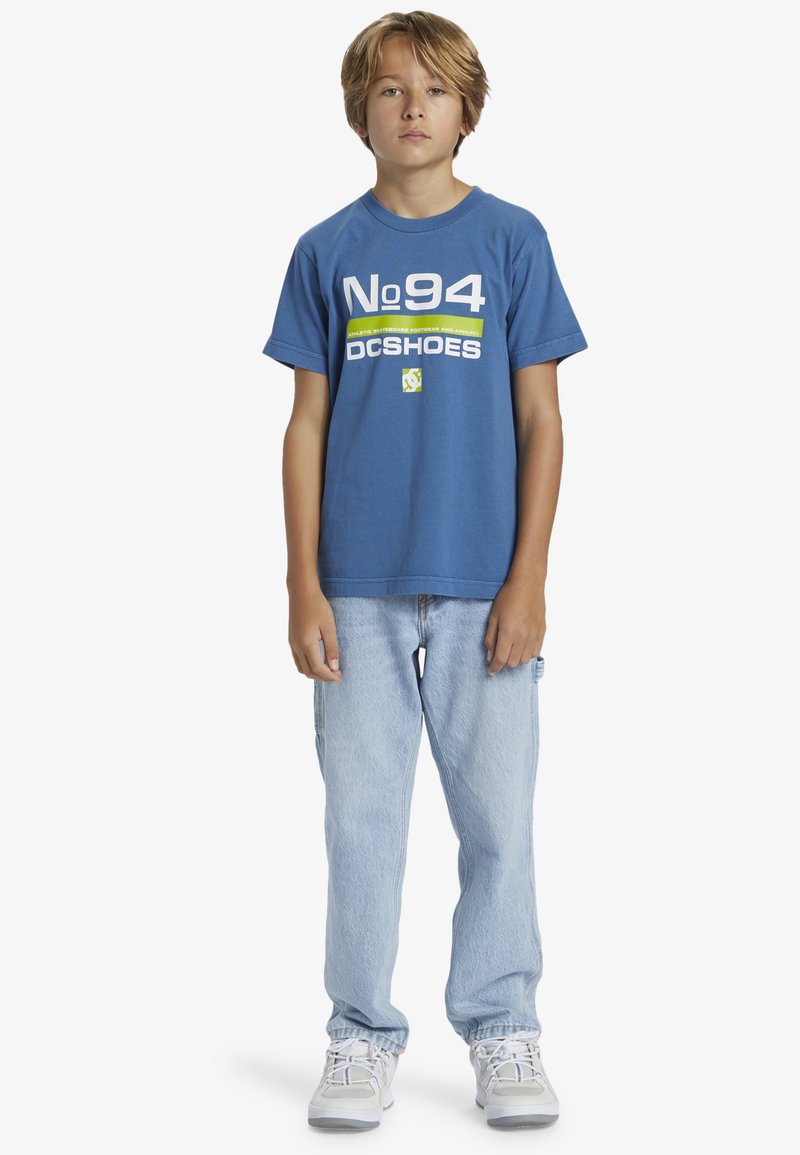 Modrá bavlněná tričko s bílým a zeleným potiskem s nápisem "No94 DC Shoes," v kombinaci se světle modrými džíny a bílými sportovními botami.