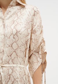 Blouse à motif python en crème et marron, avec un col à revers, des boutons sur le devant, et des manches resserrées avec des liens. Tissu à texture lisse.