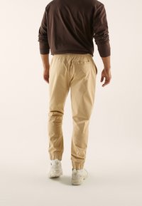 Beżowe spodnie dresowe zwężane, z elastycznym pasem i mankietami, w zestawie z brązową koszulą z długim rękawem i białymi trampkami. Widok z tyłu.