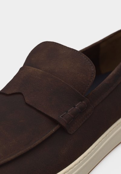 Mocassin à enfiler en daim marron foncé avec détail de sangle cousue et semelle en caoutchouc blanche sur fond blanc.