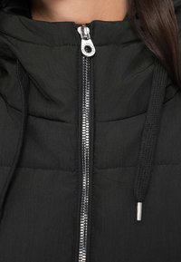 Svart boblejakke med glidelås foran, laget av et strukturert stoff, med hette og snorer. Skinnende glidelåshardware kompletterer designet.