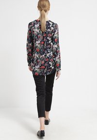 Blomsterbluse med en mørk marineblå baggrund, prydet med røde og grønne blomster, lange ærmer og en løs pasform. Sorte bukser og flade sko.