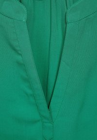 Primo piano di un tessuto verde con scollo a V profondo e cuciture visibili lungo i bordi.