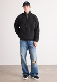 Tânăr îmbrăcat cu un fleece negru cu fermoar, blugi rupți albaștri, adidași negri și albi și o șapcă neagră, pe un fundal alb simplu.