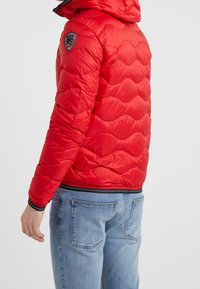 Osoba oblečená v červené prošívané bundě s černým lemem a logem na rukávu, v kombinaci se světle modrými džínami na jednobarevném pozadí.