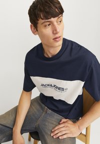 Marineblå bomulds t-shirt med en hvid vandret stribe, der har "JACK & JONES ESSENTIALS WORLDWIDE" trykt i mørkeblåt. Rund halsudskæring.