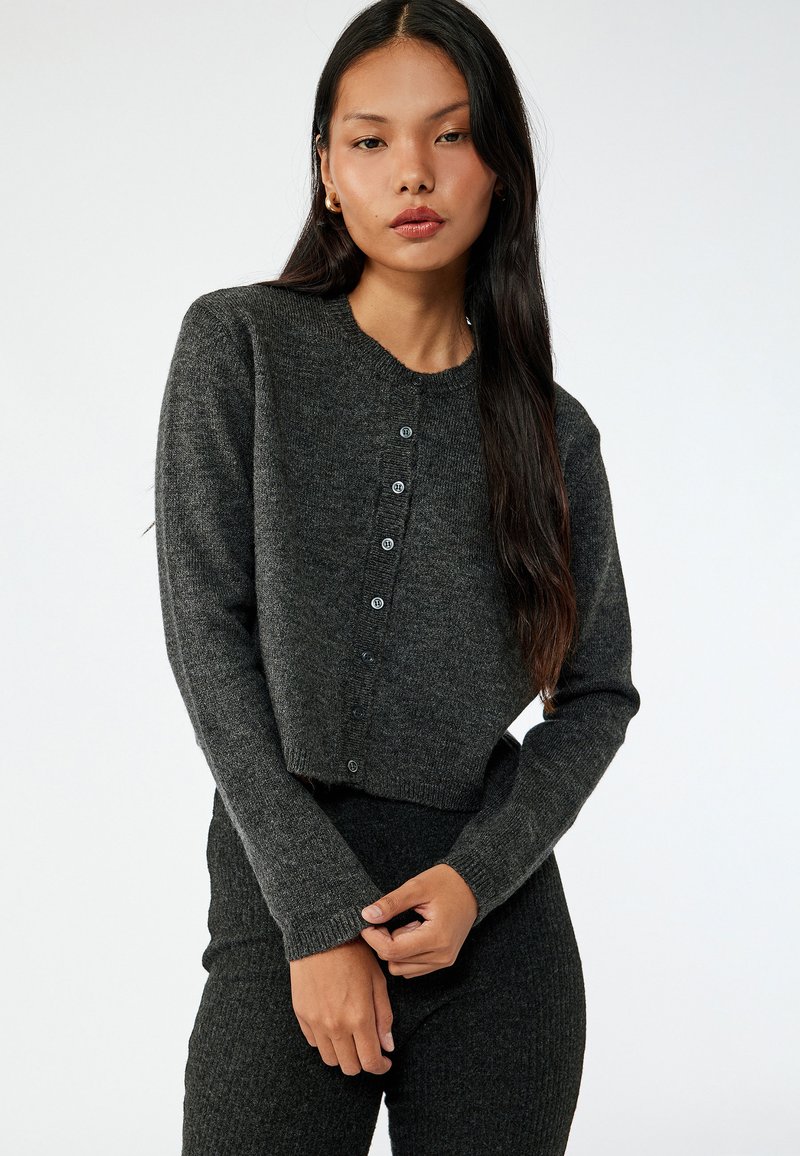 Grau geschnittene Strickjacke aus gestricktem Stoff, mit Rundhalsausschnitt und sechs Knopfverschlüssen vorne. Lange Ärmel, strukturierte Oberfläche.