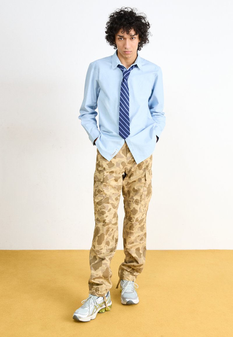 Chemise bleu clair à boutons, cravate rayée marine, pantalon cargo beige camouflage et baskets argentées avec accents verts, se tenant sur un sol jaune.