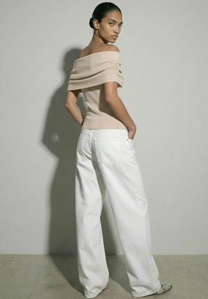 Donna con i capelli raccolti indossa un top beige scollato sulle spalle e pantaloni bianchi a gamba larga, in piedi contro una parete grigio chiaro uniforme.