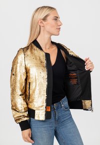 Donna con lunghi capelli biondi indossa una giacca bomber dorata con paillettes sopra una canotta nera e jeans blu, tenendo la giacca aperta per mostrare la fodera nera.