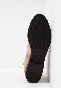 Bottine en daim beige clair découpée, avec une semelle en caoutchouc noire texturée présentant un motif géométrique et un détail de fermeture éclair sur le côté.