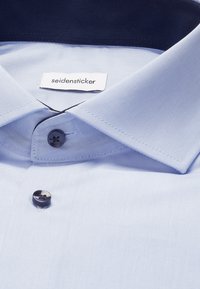 Jasnoniebieska koszula z ciemnymi guzikami i złożonym kołnierzem, na którym widać białą metkę marki z napisem "seidensticker."