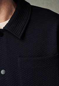 Tmavě modrá prošívaná bunda s klasickým límcem, zapínáním na knoflíky a malou přední kapsou s jemnou texturou.