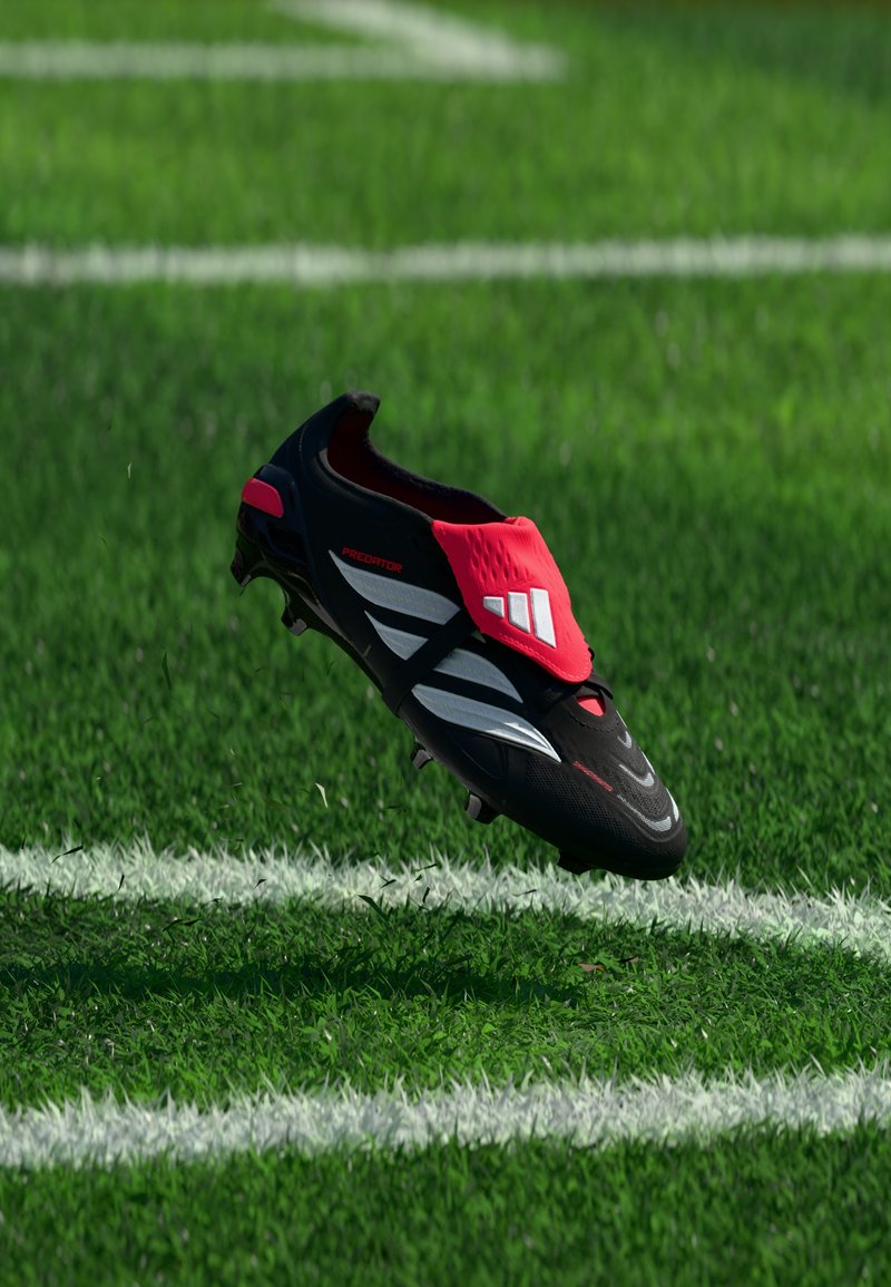 Μαύρο και κόκκινο ποδοσφαιρικό παπούτσι Adidas Predator στον αέρα πάνω από πράσινο τεχνητό χλοοτάπητα με λευκές γραμμές γηπέδου.