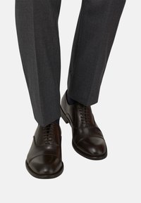 Scarpe eleganti in pelle marrone con punta tonda, dettagli a punta chiusa e lacci, abbinate a pantaloni grigi su misura e calze scure.