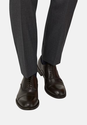 Czarne skórzane buty eleganckie z zaokrąglonym noskiem, detalami w kształcie czubka i sznurówkami, zestawione z szarymi, dopasowanymi spodniami i ciemnymi skarpetami.