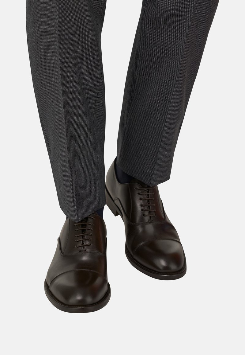 Scarpe eleganti in pelle marrone con punta tonda, dettagli a punta chiusa e lacci, abbinate a pantaloni grigi su misura e calze scure.