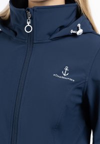 Marinblå jacka med hög krage, dragkedjestängning och vit broderad ankarlogo. Slätt, vattenavvisande material med detaljer av dragsko i huva.