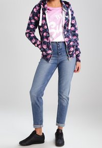 Felpa con zip a fondo navy e motivo floreale, con lacci bianchi, sopra una maglietta grafica rosa. Jeans a vita alta azzurri e scarpe da ginnastica nere.