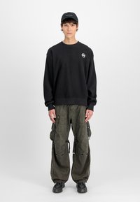 Man wearing a black cap, oversized black textured sweatshirt, loose dark cargo pants, and black sneakers, standing against a plain white background.
