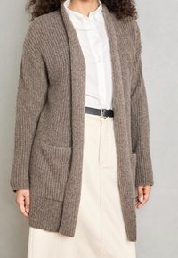 Brun ribbad cardigan i texturerat tyg, med två framsidor fickor och en öppen design, buren över en vit helknappad skjorta.