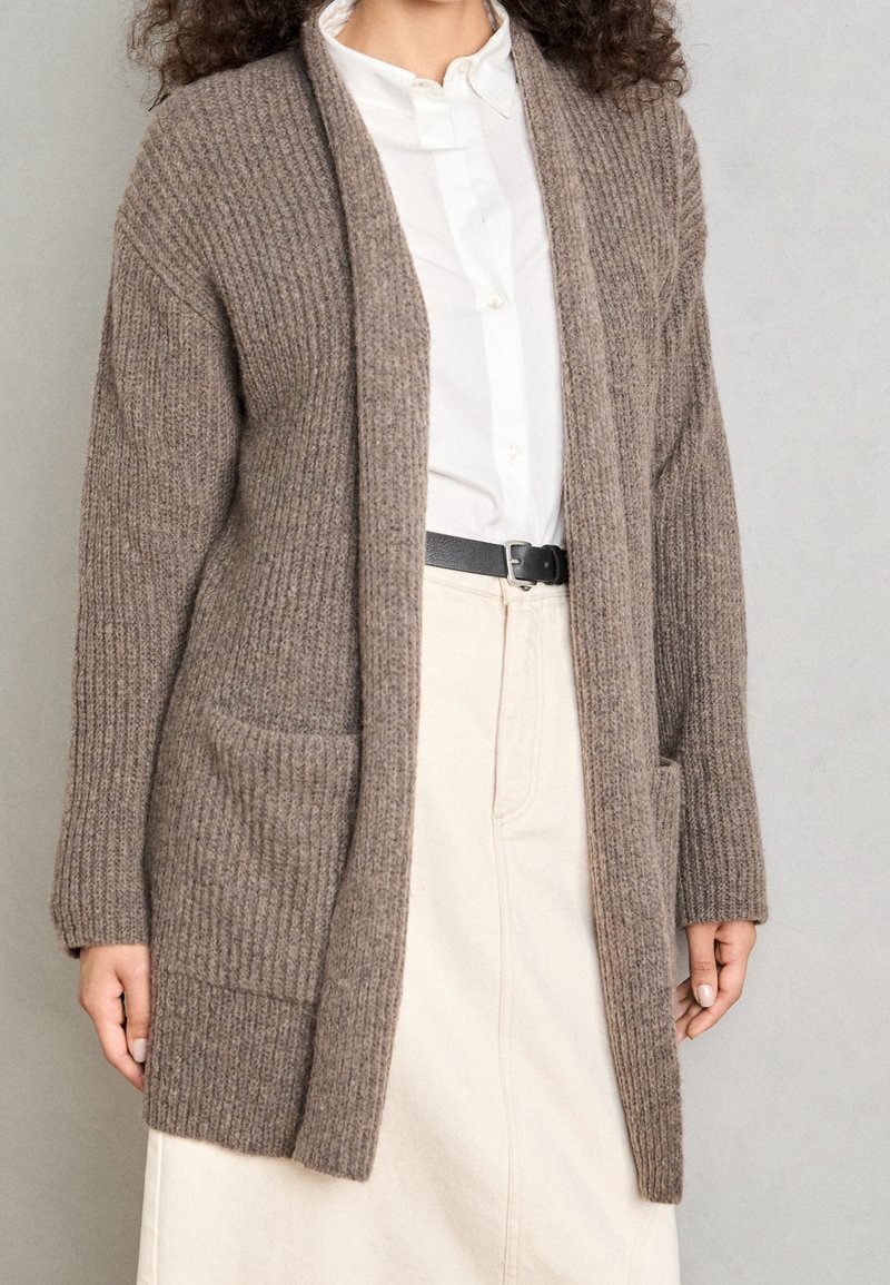 Brun ribbad cardigan i texturerat tyg, med två framsidor fickor och en öppen design, buren över en vit helknappad skjorta.