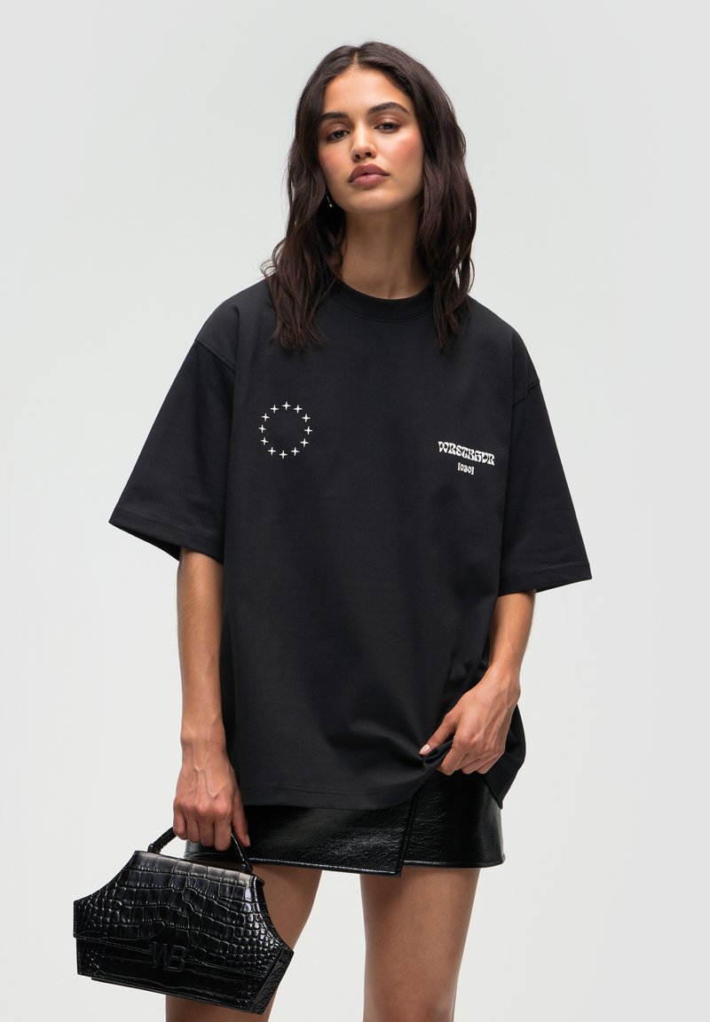 Zwart oversized t-shirt met witte grafische elementen, gecombineerd met een glanzende zwarte handtas met krokodillenstructuur en een glanzende zwarte minirok.