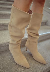 Botas de rodilla de ante beige de tacón alto con puntera afilada y parte superior suave y fluida. Presenta un diseño elegante y ajustado con tacón delgado.