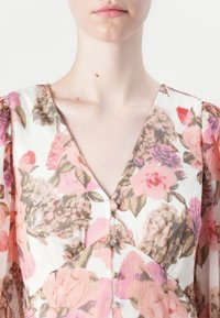 Primer plano del cuello y la parte superior del pecho de una persona de piel clara que lleva una blusa floral translúcida con flores rosas, moradas y marrones y botones cubiertos de tela.