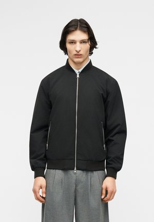 Νεαρό άτομο με σκούρα μαλλιά που φοράει μαύρο μπουφάν τύπου bomber με φερμουάρ και γκρι πλισέ παντελόνι, στέκεται μπροστά σε απλό ανοιχτό φόντο.