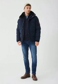 Tengerészkék pufferekedő bunda, bolyhos kapucnival, cipzáras elülső résszel és logós folttal. Sötét pulóver fölött viselve, kék farmerral és barna csizmával párosítva.