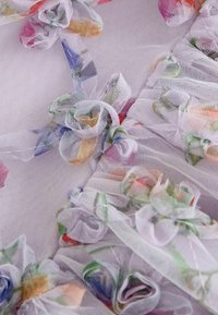 Světle fialová tkanina zdobená jemnými květinovými vzory a malými aplikacemi z tylu ve tvaru růží, s vrstvenými texturálními prvky v různých odstínech.