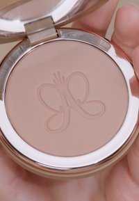 Compact con una tonalità beige chiaro, caratterizzato da un logo di una farfalla in rilievo. La confezione ha un bordo argento e un coperchio trasparente.