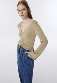 Beige gestrickter Top mit V-Ausschnitt, tailliert mit Kordelzügen, kombiniert mit lockeren blauen Jeans. Lange, ausgestellte Ärmel.