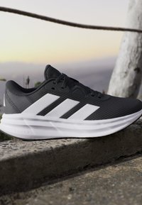 Chaussure de sport noire avec des bandes blanches et une semelle blanche épaisse posée sur un rebord en pierre avec un arrière-plan extérieur flou au coucher du soleil.