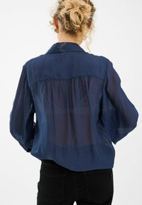 Tumedas sinine, kerge shirt, millel on läbipaistev kangas, pikad varrukad ja seljaõmbluse disain. Omab teravat kraed ja vabalt langevat lõiget.