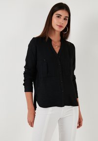 Black button-up shirt made of textured fabric, featuring long sleeves, two chest pockets, and a v-neck design. Paired with white striped pants.