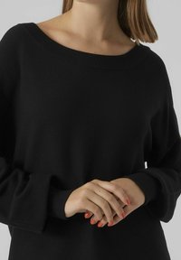 Pull noir en maille avec un large col et des manches longues. Le tissu semble doux avec des poignets côtelés, offrant une coupe décontractée.
