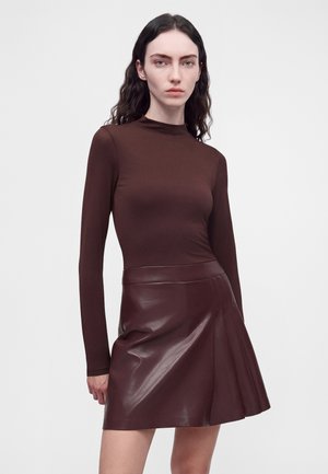 Femme aux longs cheveux foncés portant un haut marron foncé cintré à manches longues et une jupe en cuir marron foncé brillante sur un fond clair uni.