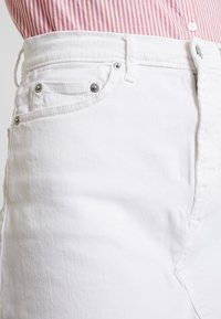 Bílé džíny s vysokým pasem, které mají přední kapsu, kovové knoflíky a mírnou texturu. Materiál je hladký a neprůhledný.