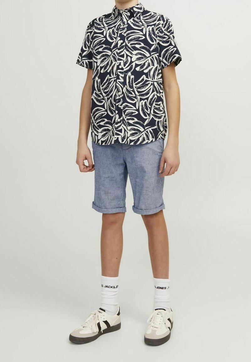 Personne portant une chemise à manches courtes imprimée de feuilles bleu marine et blanches, un short bleu clair retroussé, des chaussettes blanches et des baskets beige.