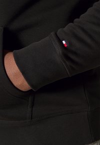 Felpa nera con texture a maglia, dotata di un sottile patch con logo nei colori rosso, bianco e blu sul polsino. Dettagli delle cuciture visibili.