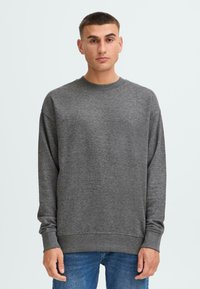 Ung mann iført en enkel, mørkegrå langermet sweatshirt og blå jeans, stående mot en lys bakgrunn, vendt fremover.