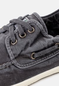 Scarpa in tela grigia con occhielli in metallo e lacci grigi. Presenta una suola in gomma bianca e cuciture a contrasto. Texture morbida.