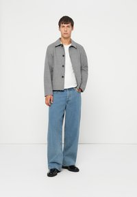 Grå rutete jakke med svarte knapper, iført over en hvit skjorte. Kombinert med lyseblå vide jeans og svarte sko.